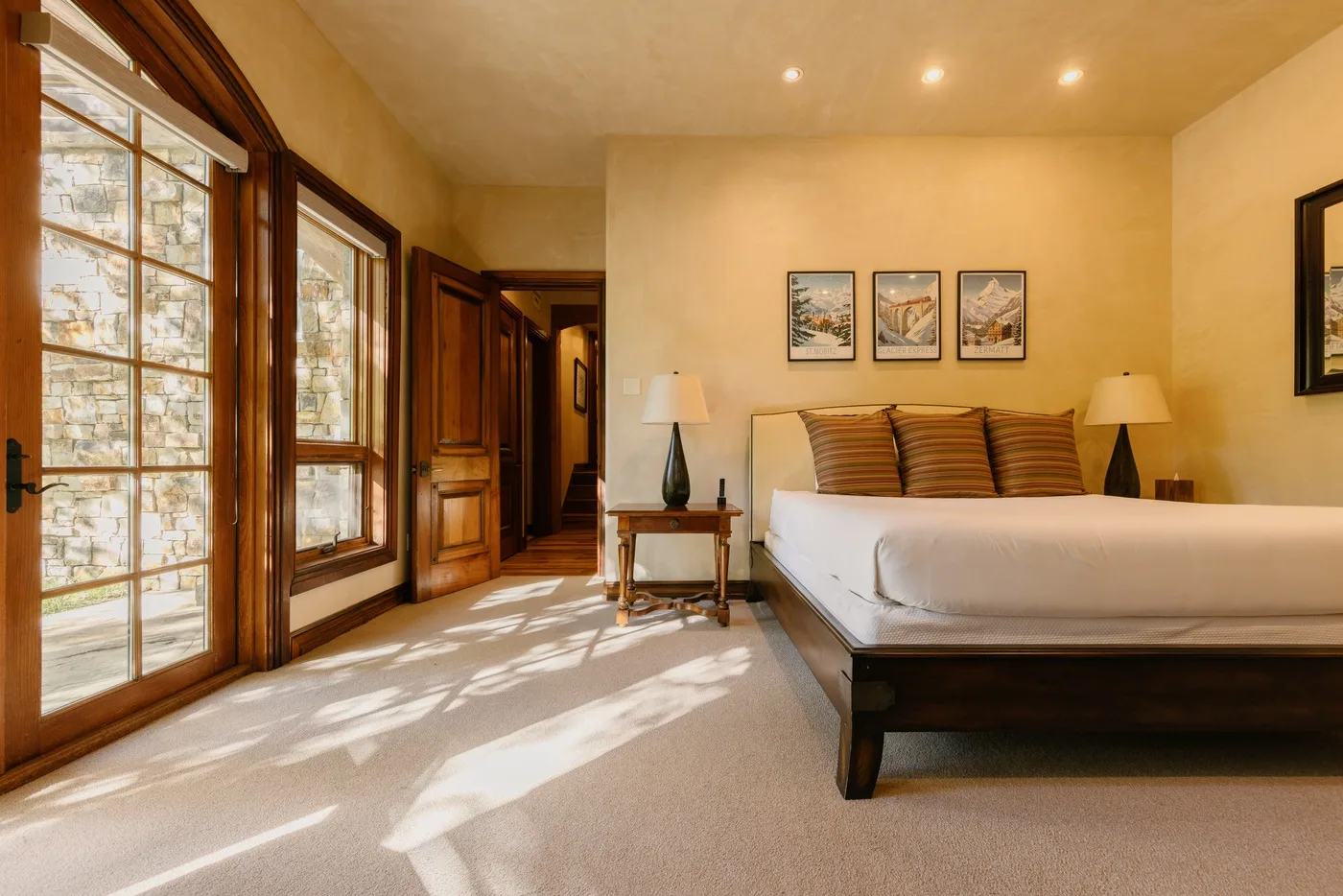 A bedroom with a bed, framed pictures, and tall windows letting in sunlight.