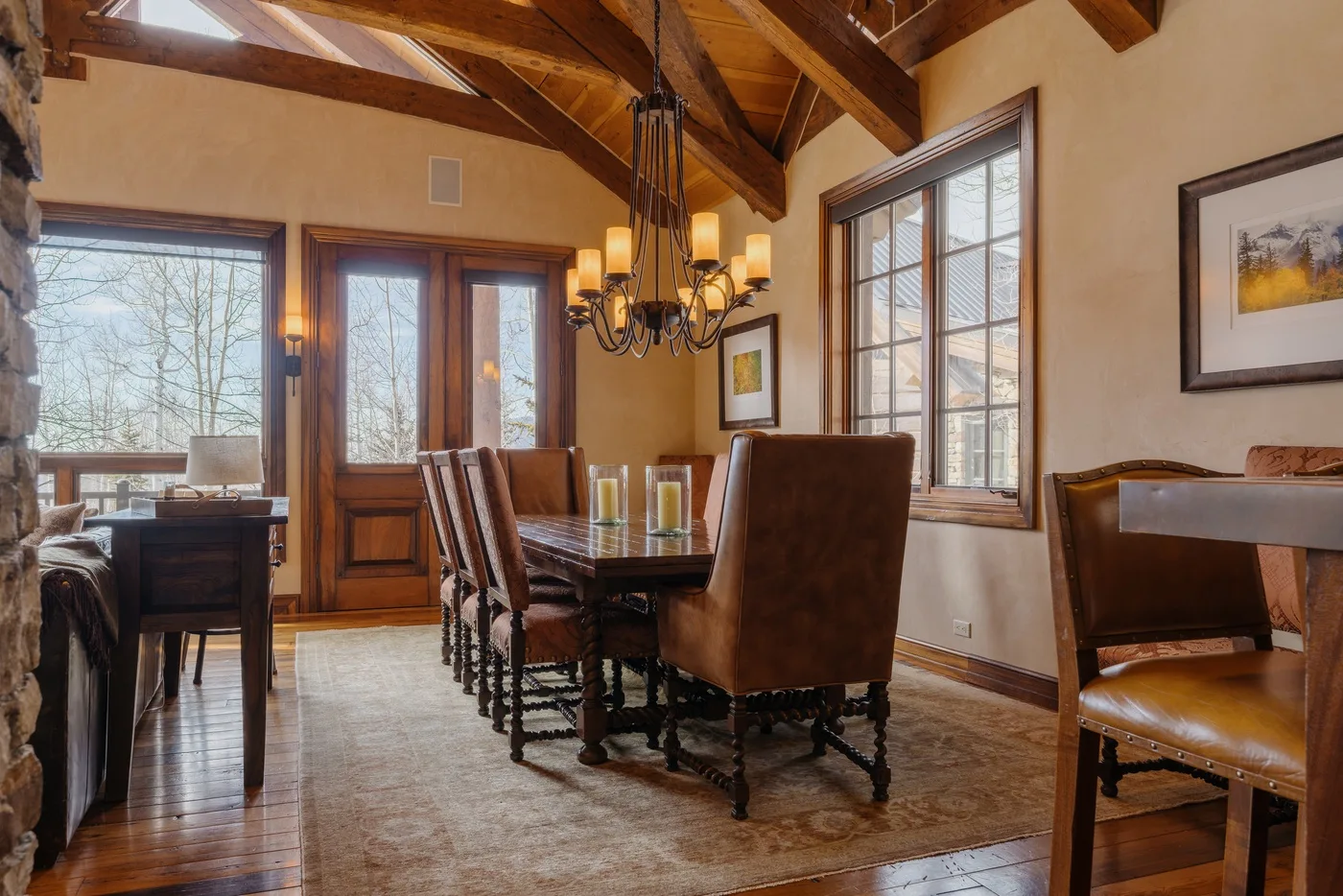 A dining room with a long table and large windows beneath exposed wooden beams.