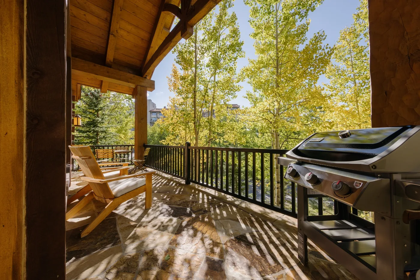 A covered wooden deck has outdoor seating, a grill, and views of leafy trees.