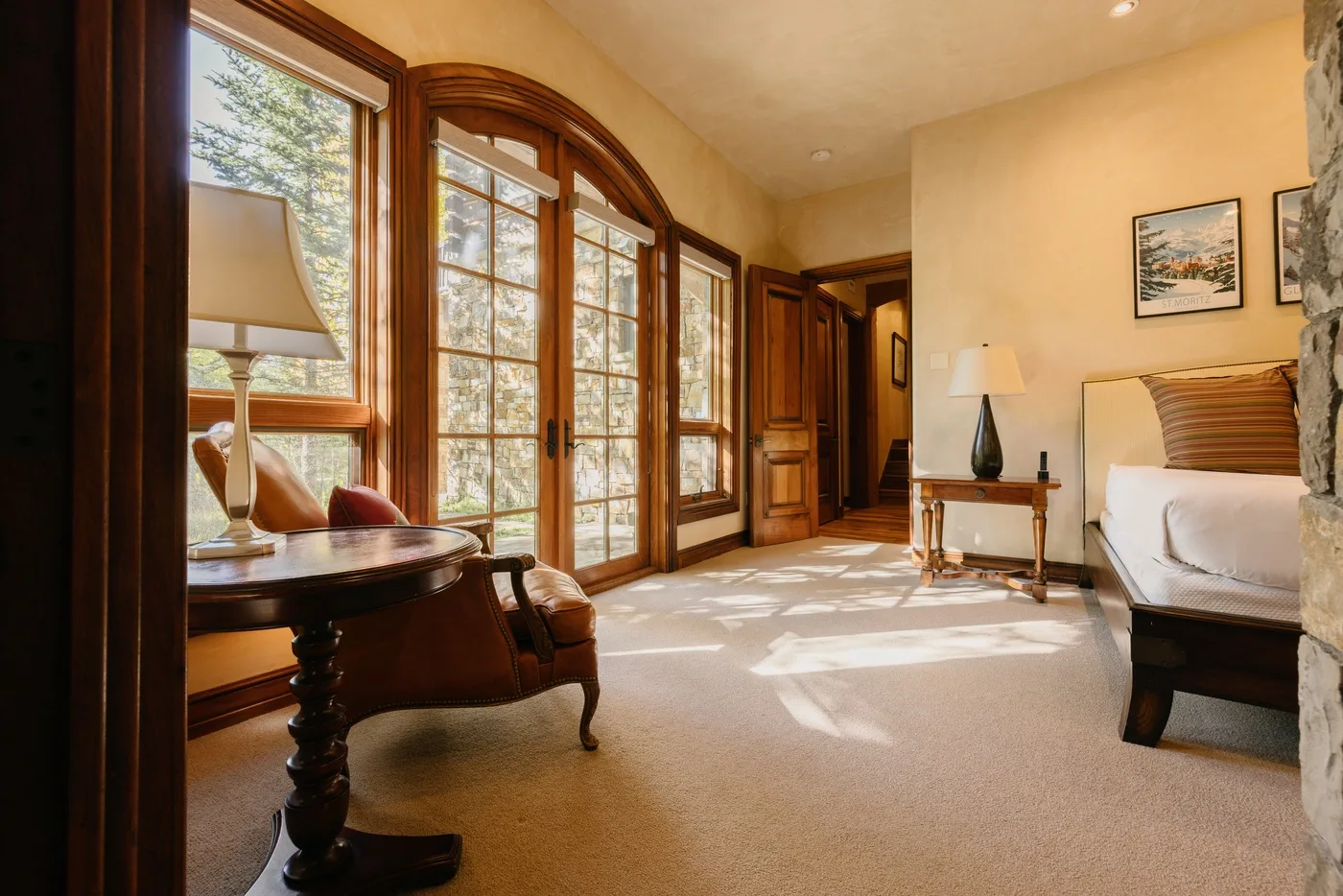 A bedroom with a bed, wooden desk, and tall windows letting in daylight.
