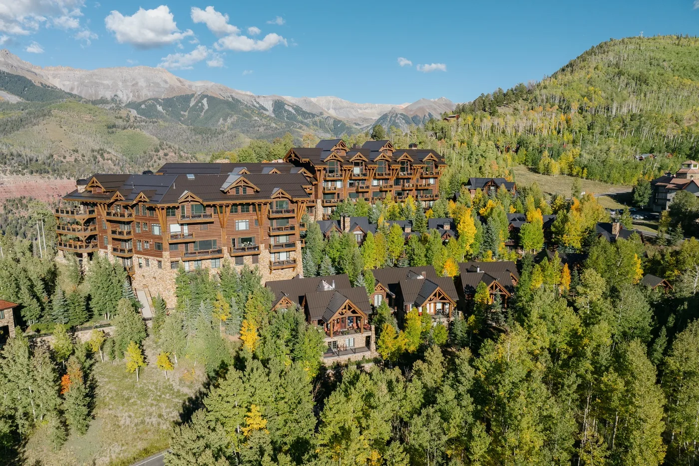 A cluster of lodge-style buildings sits among forested hills beneath a mountain range.