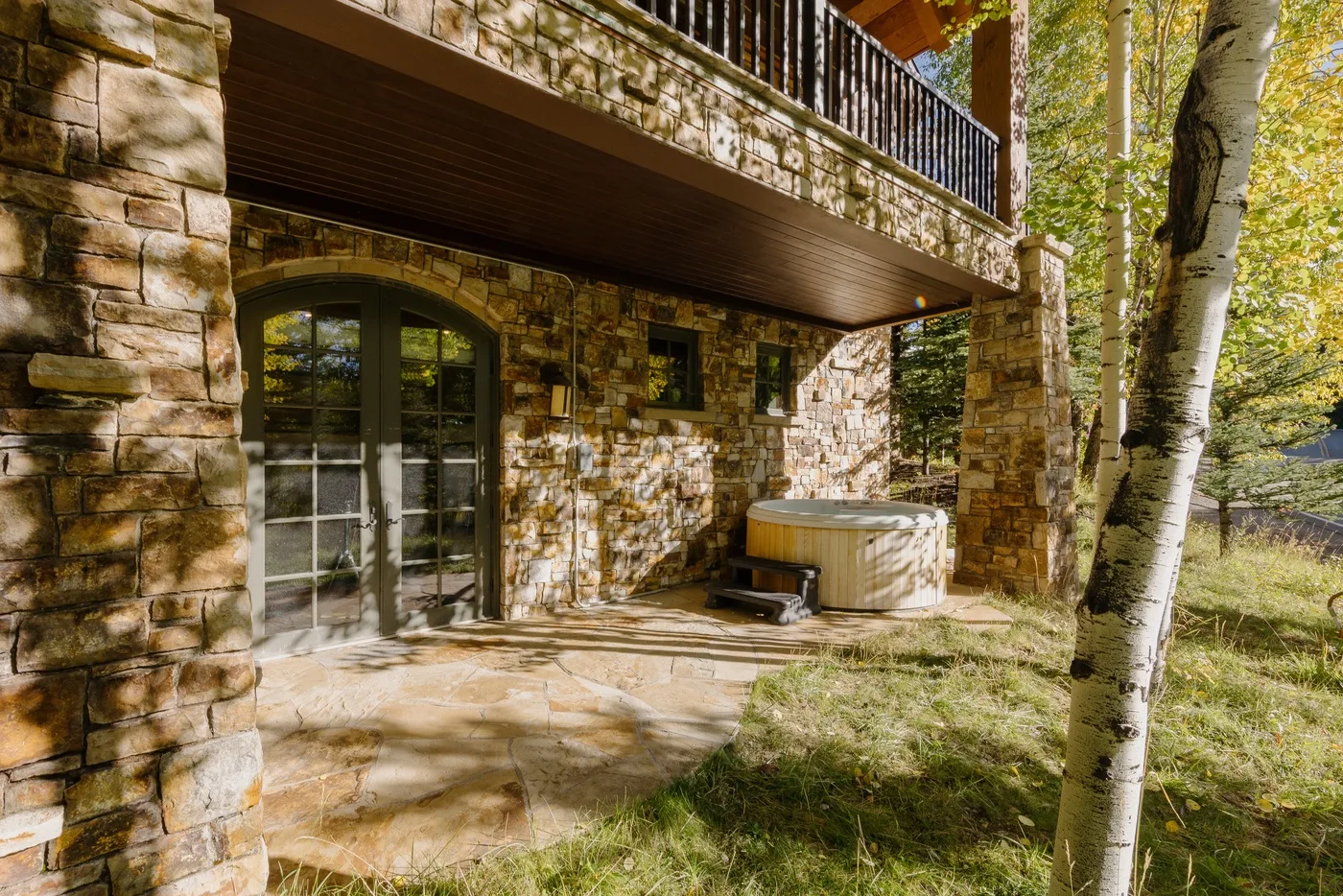 A stone patio beneath a balcony features glass doors and an outdoor hot tub.