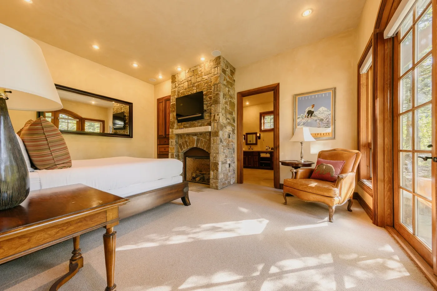 A bedroom with a bed, stone fireplace, and armchair beside tall windows.