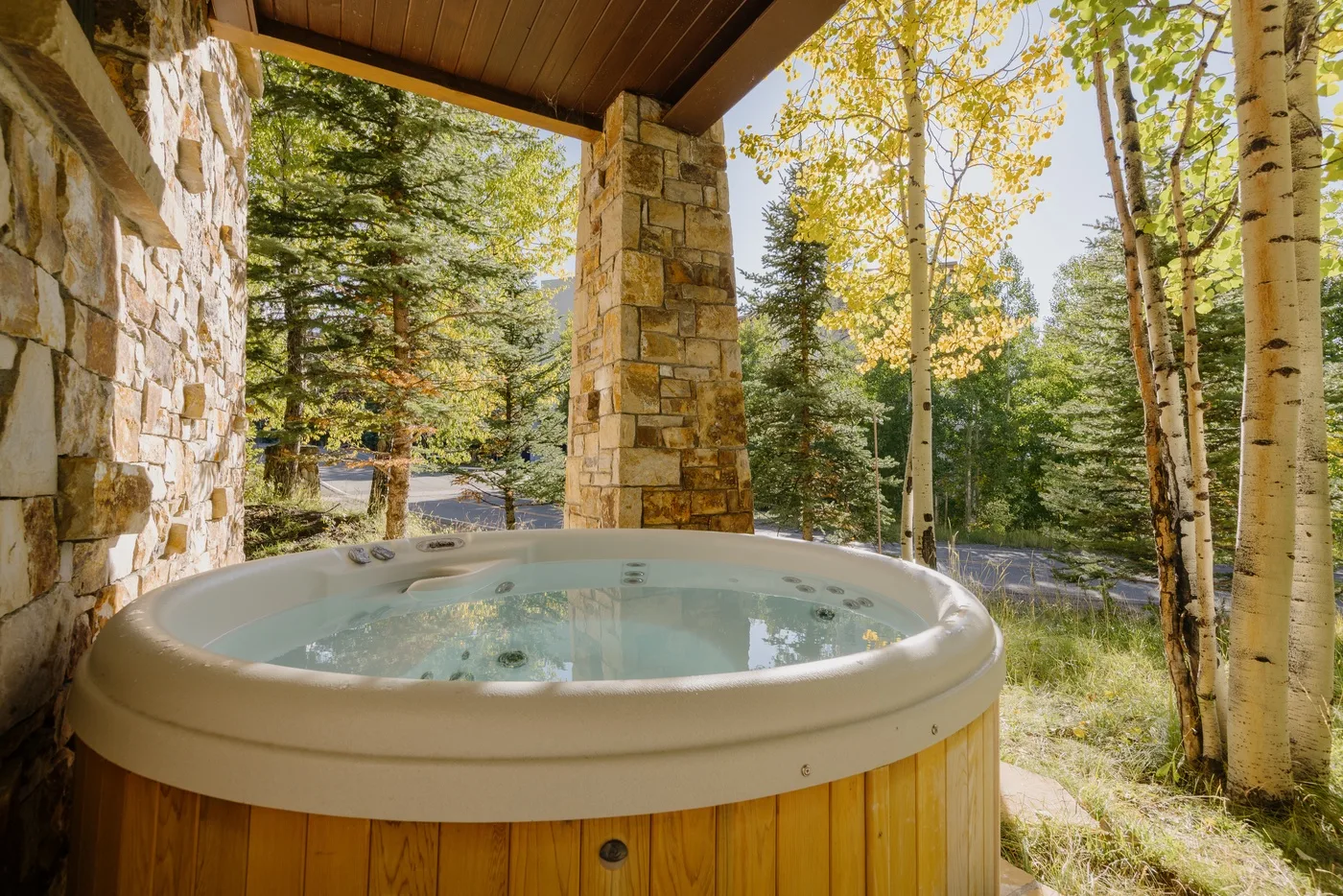 A round hot tub sits on a covered stone patio beside a wooded landscape.