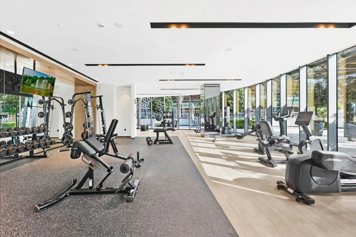 A bright gym with exercise equipment and cardio machines beside large windows.