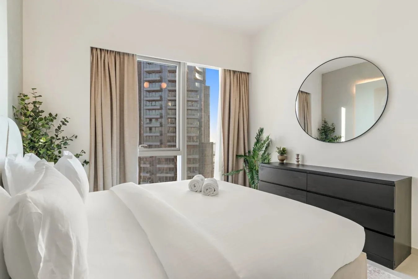 A bright bedroom with a neatly made bed, tall windows, and a round wall mirror.