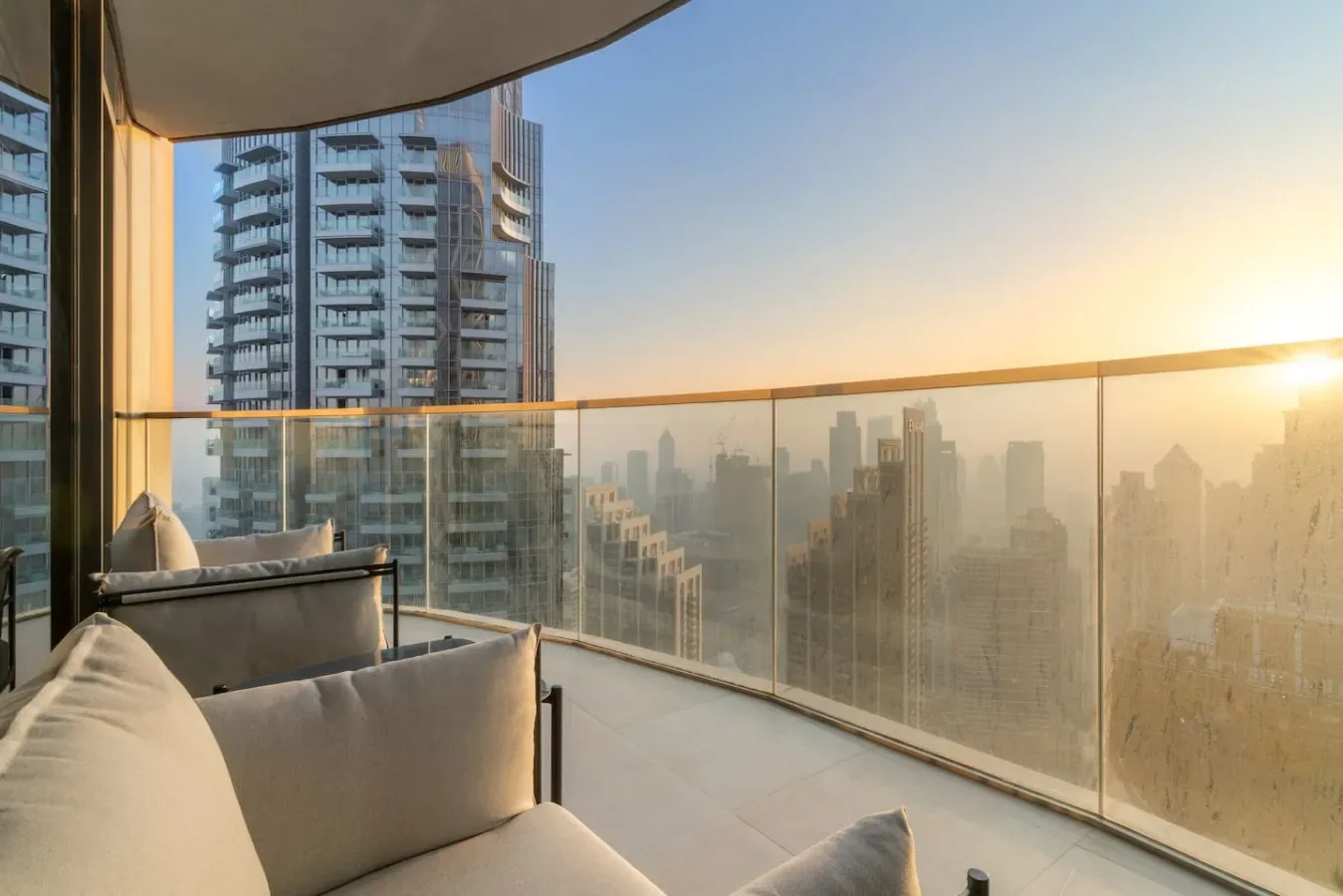A balcony with cushioned seating overlooks a city skyline at sunset.