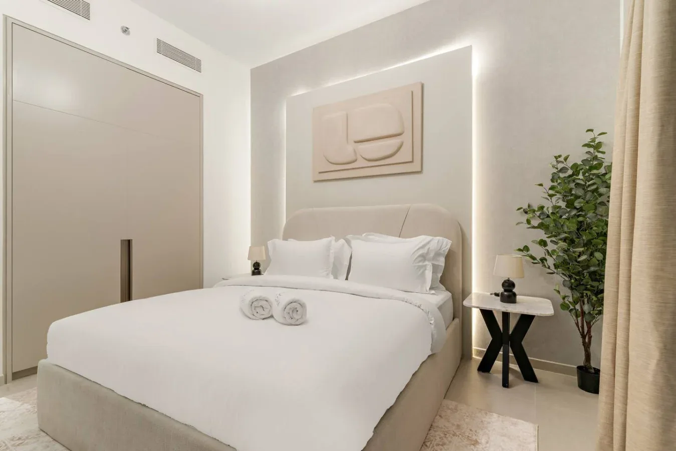 A bright bedroom with white bedding, a bedside table, and a leafy plant.