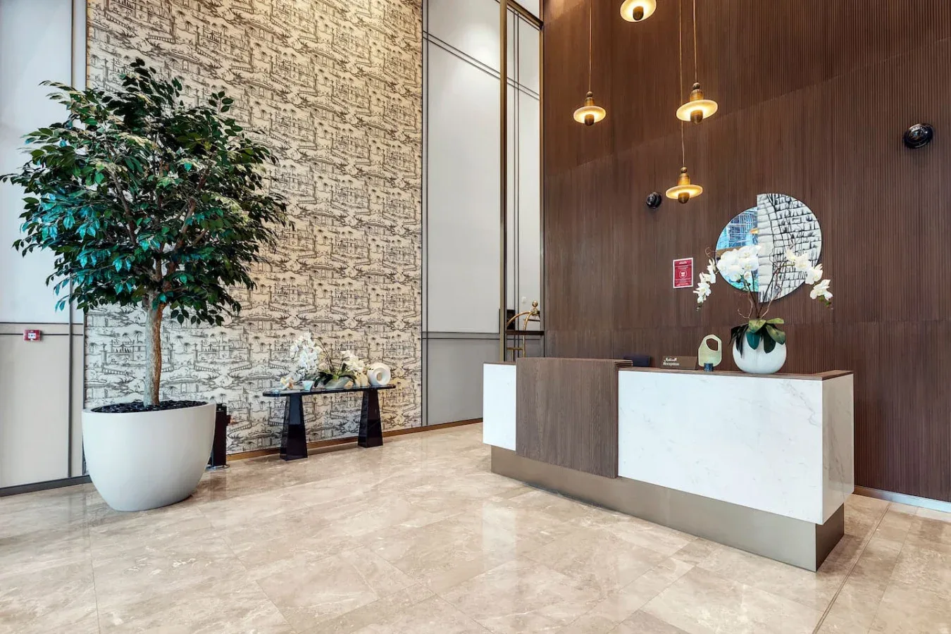 A hotel reception area with a front desk, pendant lights, and a potted tree.