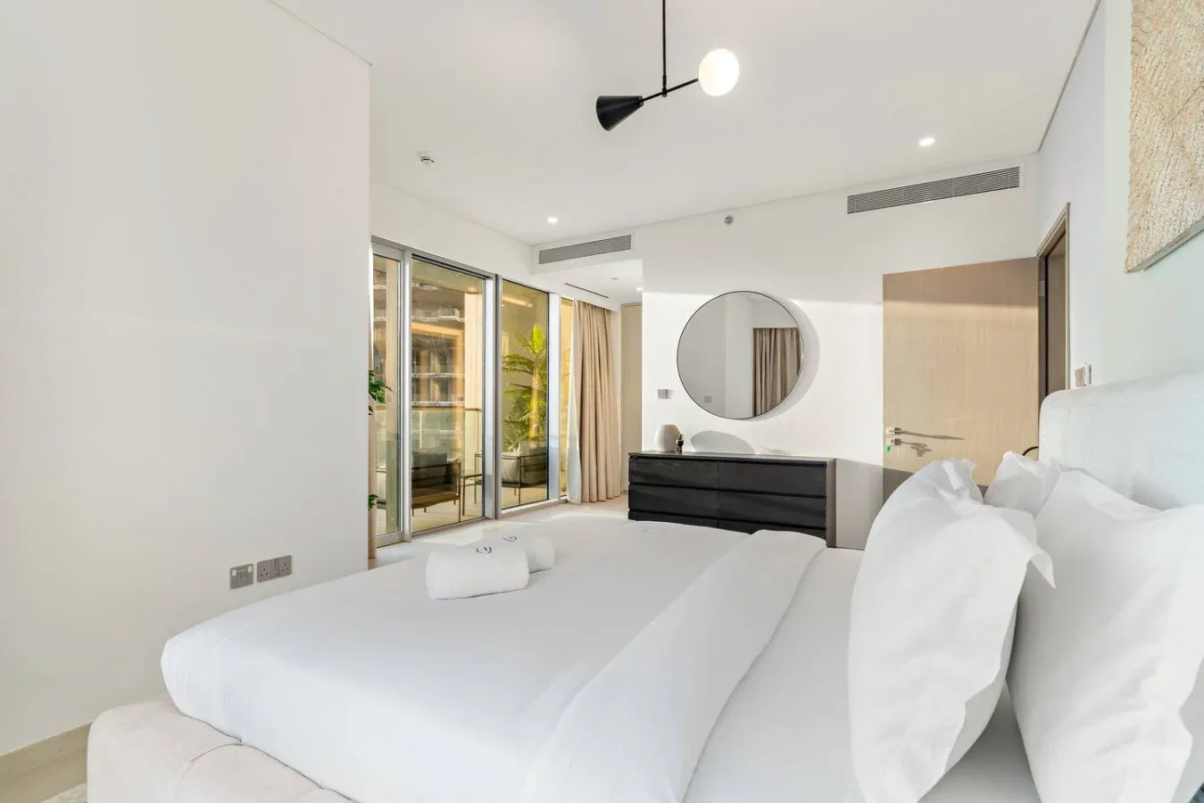 A bright bedroom with a large bed, dark dresser, and round wall mirror.