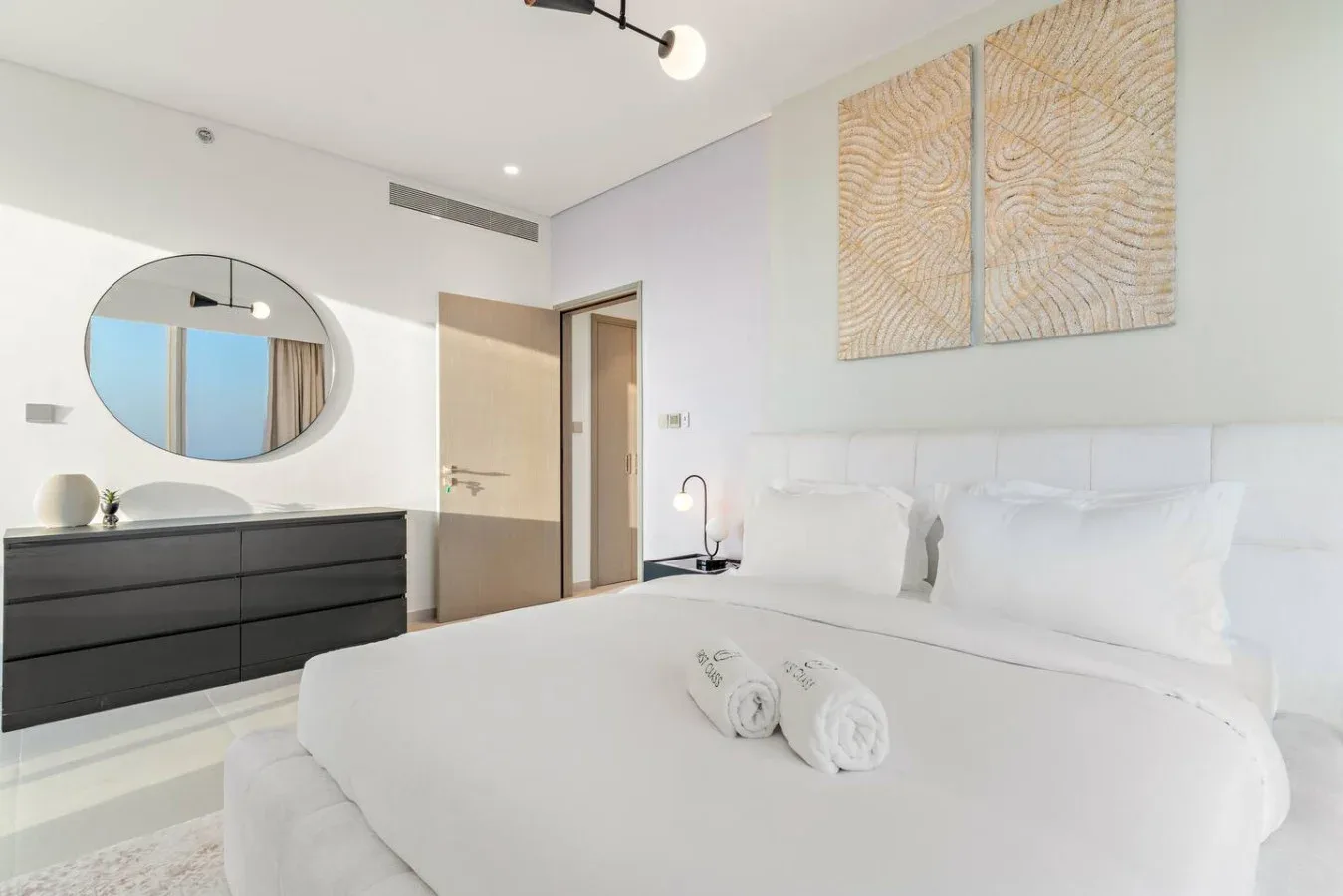 A bright bedroom with a white bed, dark dresser, and oval wall mirror.