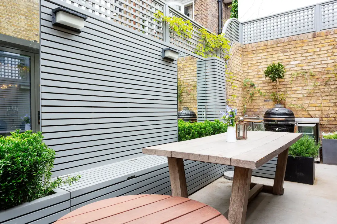 Una zona exterior pavimentada tiene una mesa de madera junto a paredes de ladrillo y vegetación.