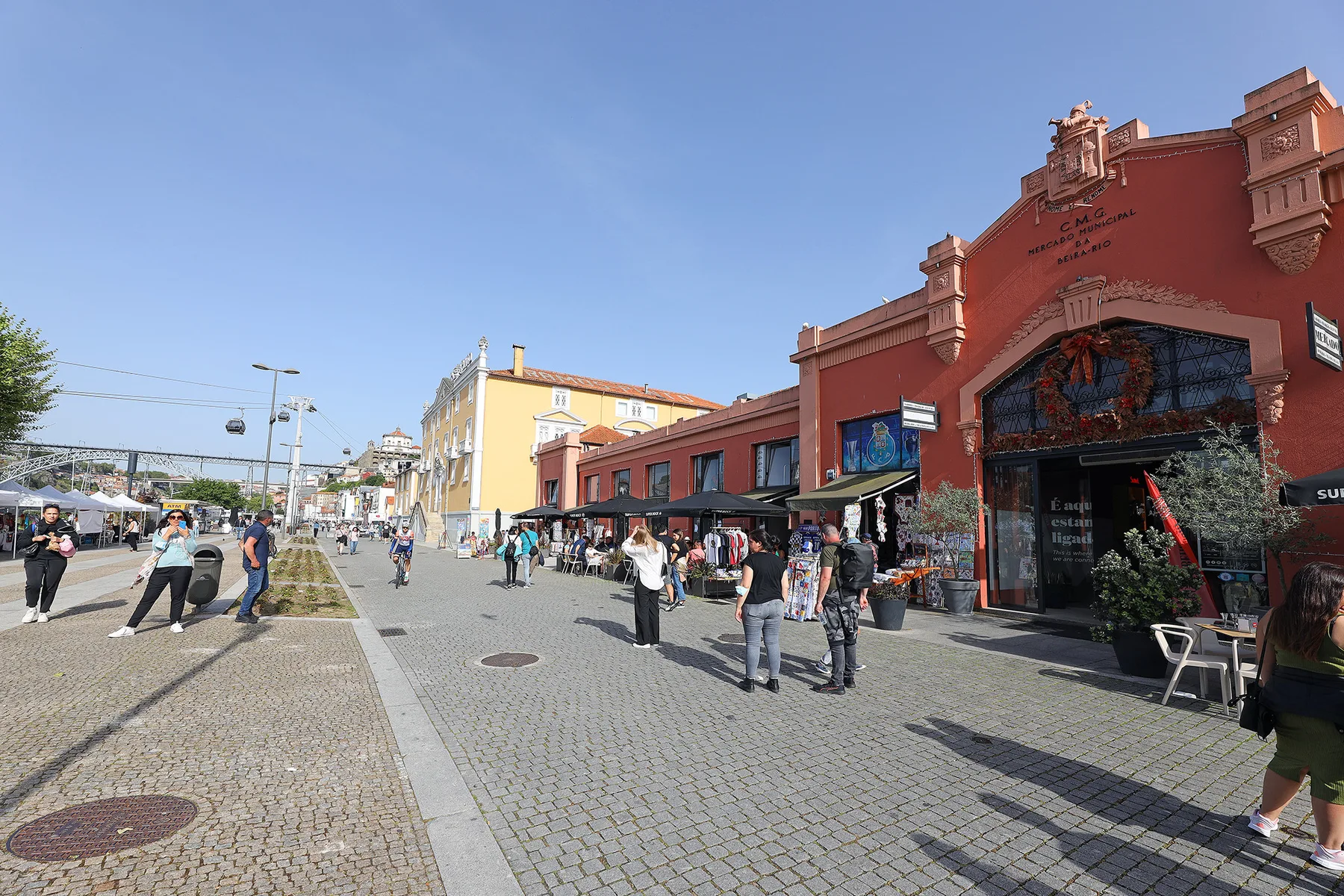 A busy pedestrian street with people walking past colorful storefronts under a clear sky.