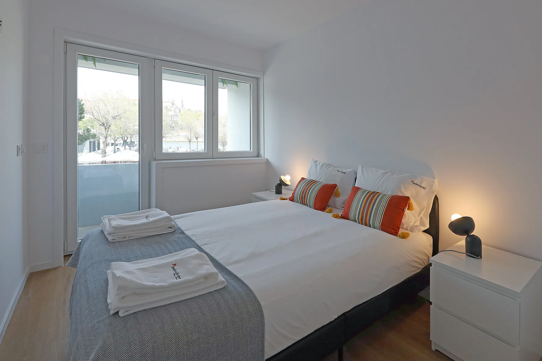 A bright bedroom with white bedding, colorful pillows, and a large window.