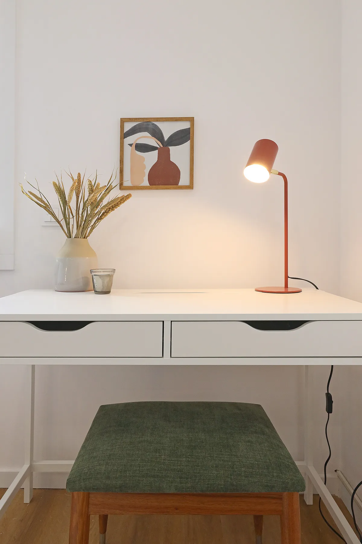 A desk with a green upholstered stool, a lit lamp, and framed artwork.