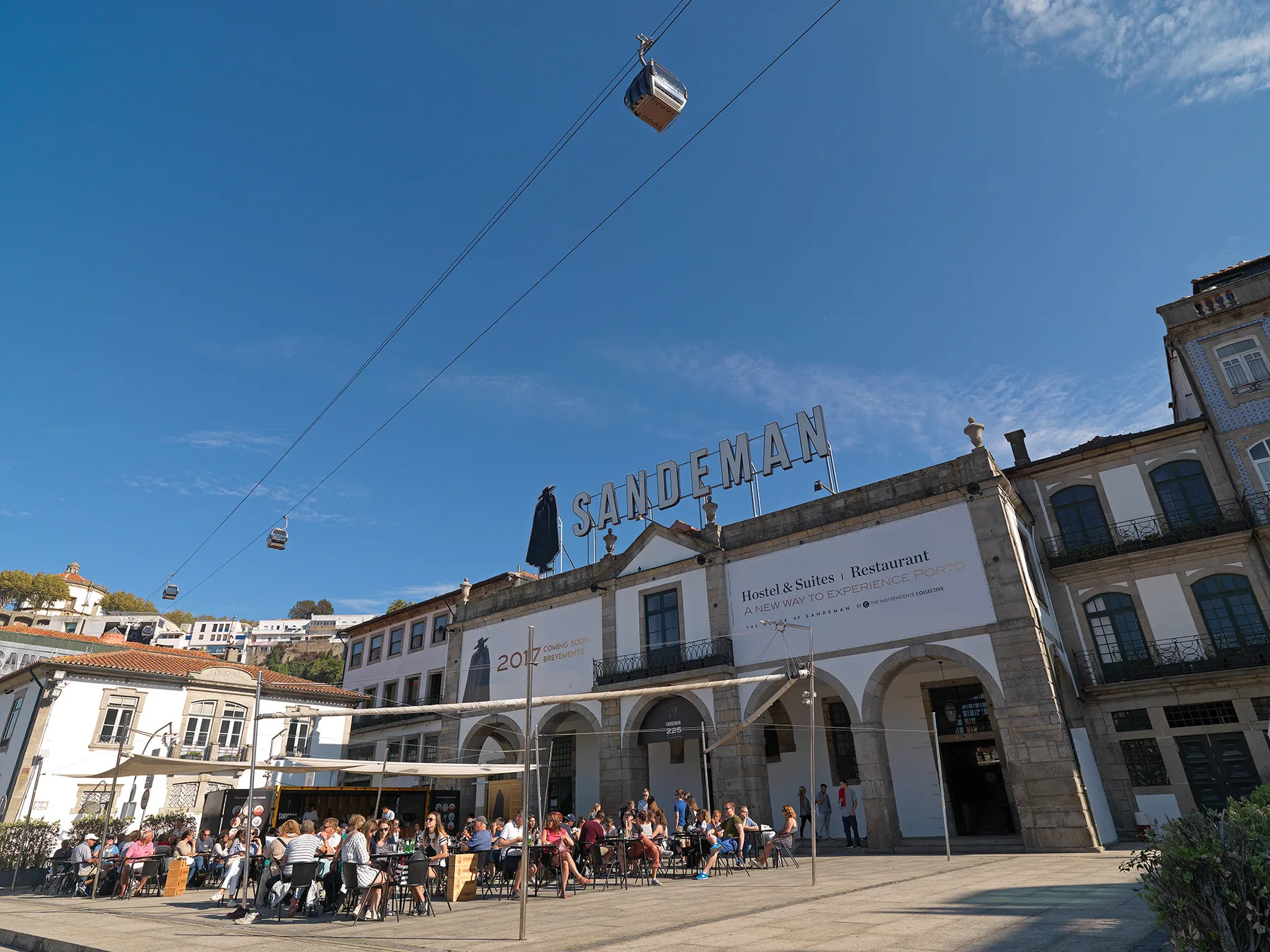 A busy outdoor café terrace sits in front of a large historic-looking building beneath a blue sky.