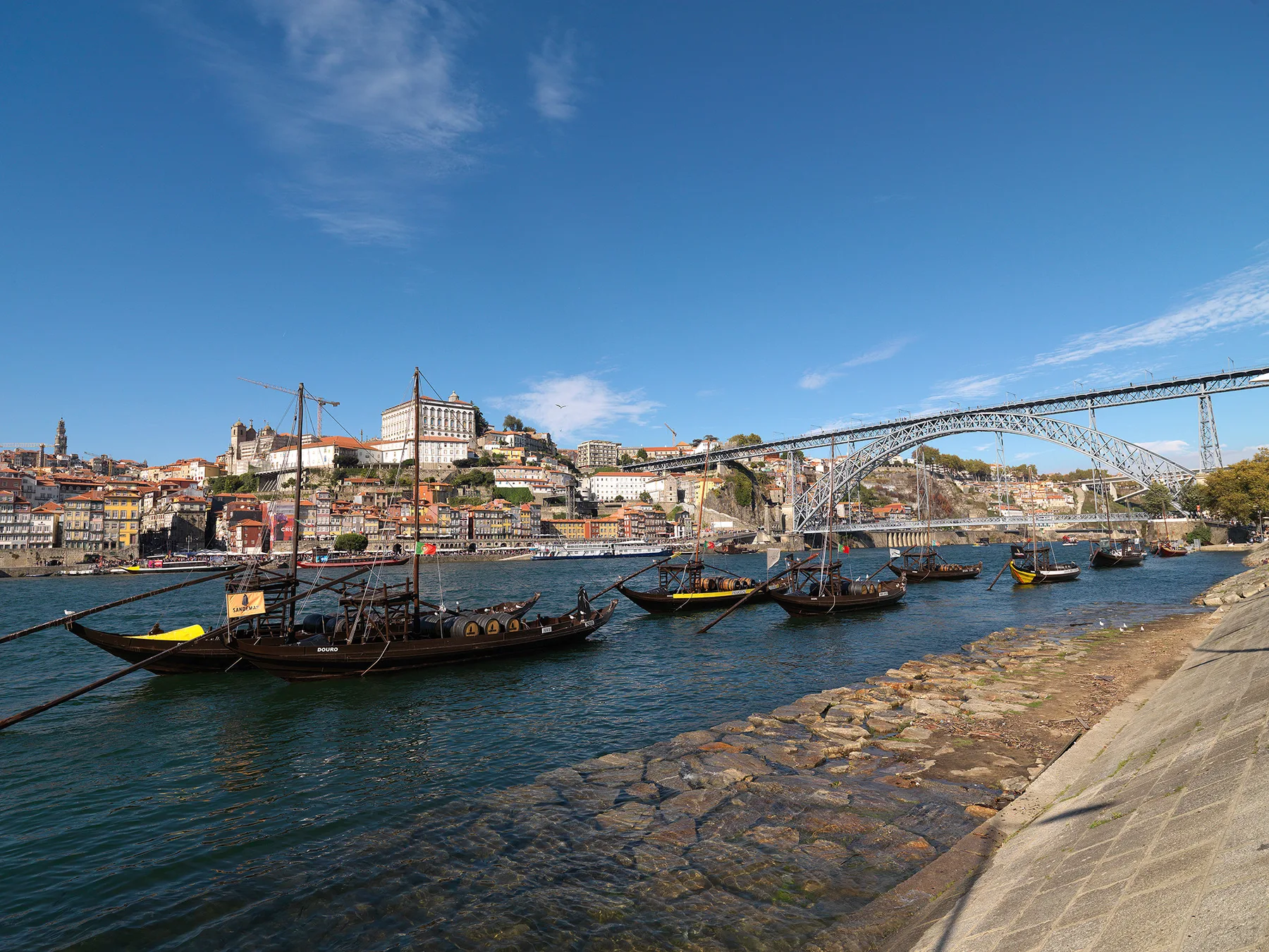 A riverfront scene with traditional boats, hillside buildings, and a large metal bridge.