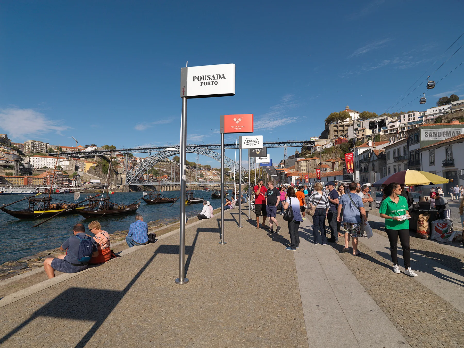 A busy riverside promenade in Porto, with pedestrians and boats along the water.