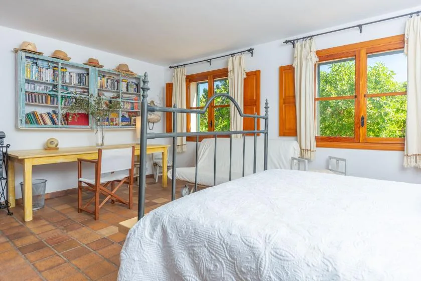 A bedroom with a large bed, a desk, and bookshelves beside the windows.