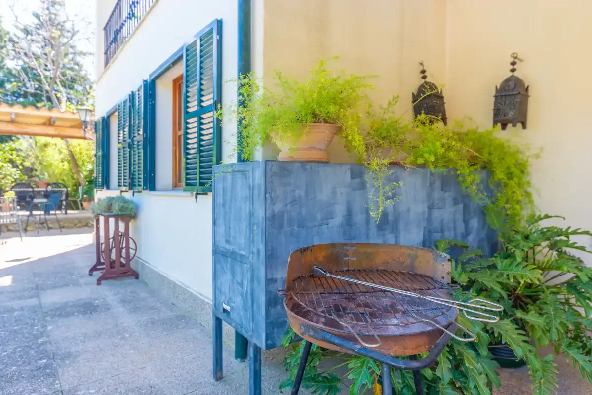 An outdoor patio with a round grill beside a blue planter and leafy plants.