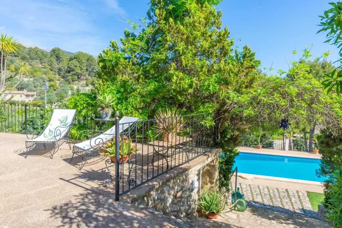A sunny outdoor patio with seating beside a swimming pool and leafy trees.