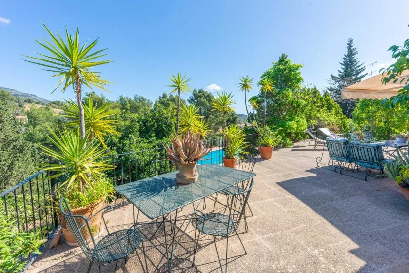 A sunny outdoor terrace has metal seating, potted plants, and a pool nearby.