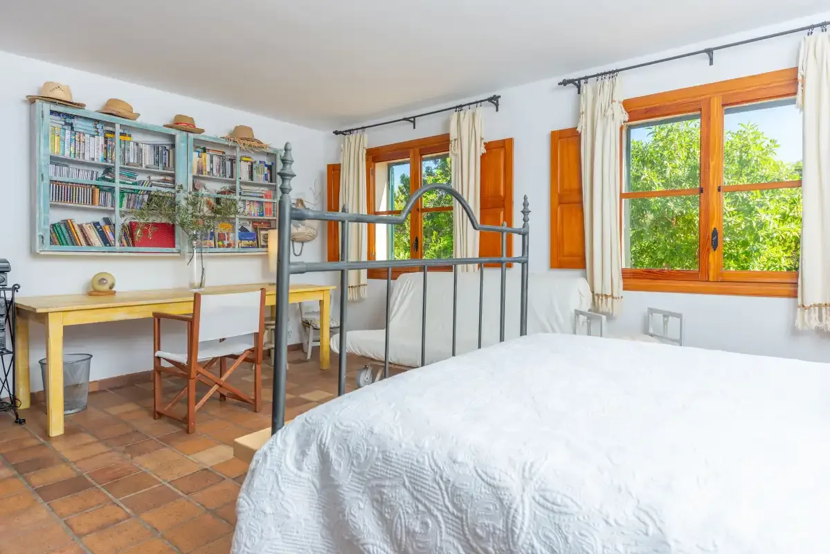 A bedroom with a large bed, a desk, and bookshelves beside the windows.