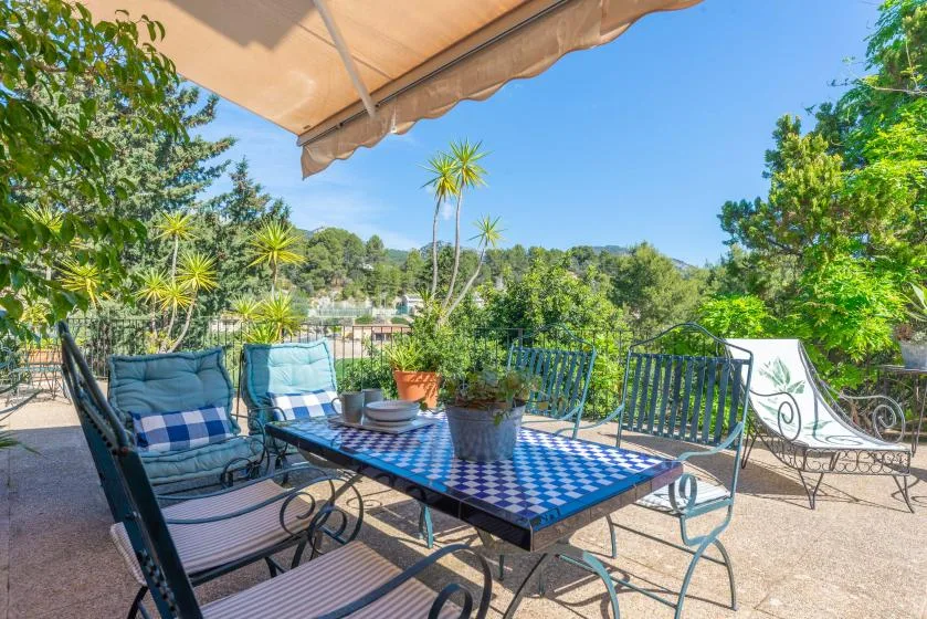 A sunny outdoor terrace has a checkered table, cushioned seating, and leafy views.