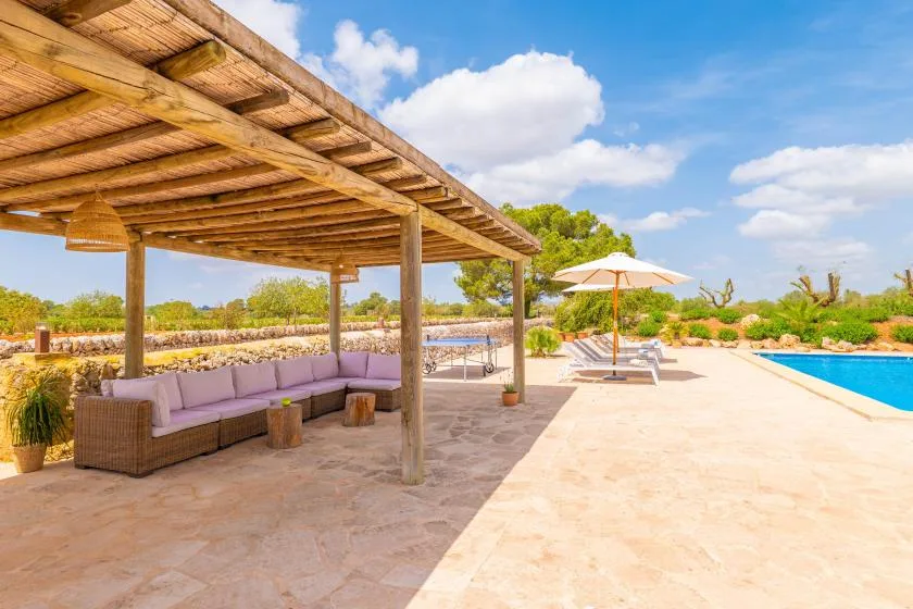 Un patio soleado junto a la piscina tiene asientos a la sombra de una pérgola de madera.