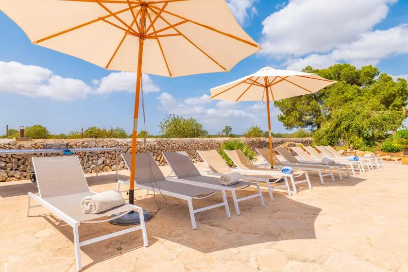 Un patio soleado junto a la piscina tiene filas de tumbonas bajo grandes sombrillas.