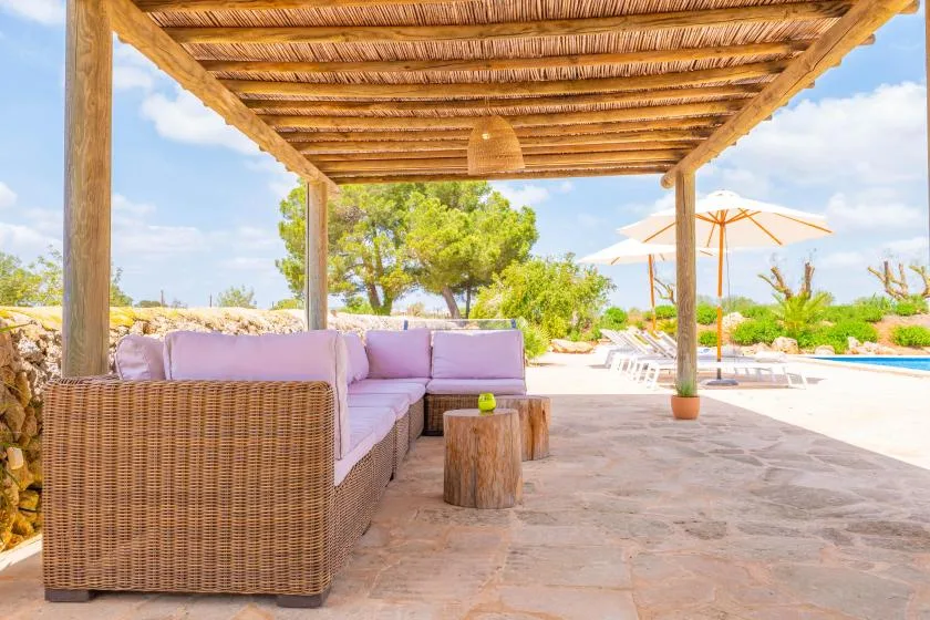 Una zona de estar exterior a la sombra de una pérgola de madera da a un patio soleado.