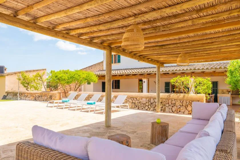 Una zona de estar al aire libre queda bajo una pérgola de madera junto a un muro de piedra.