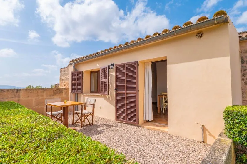 Un patio soleado junto a una casa, con asientos al aire libre y puertas con contraventanas abiertas.