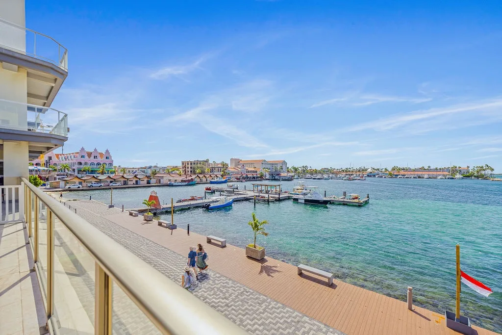 Bright seaside promenade with docked boats and colorful architecture frames a tranquil Caribbean-style marina setting beneath clear blue skies.