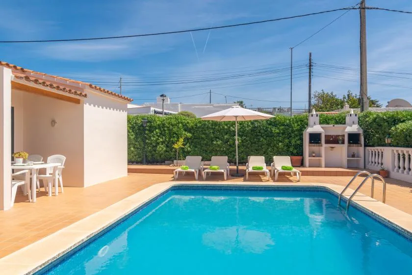 A sunny outdoor pool area with lounge seating, an umbrella, and a built-in barbecue.