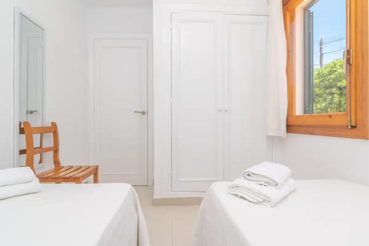 A bright bedroom with two beds, folded towels, and a wooden-framed window.