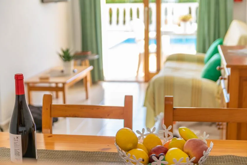 A dining table with fruit overlooks a bed and a bright balcony.