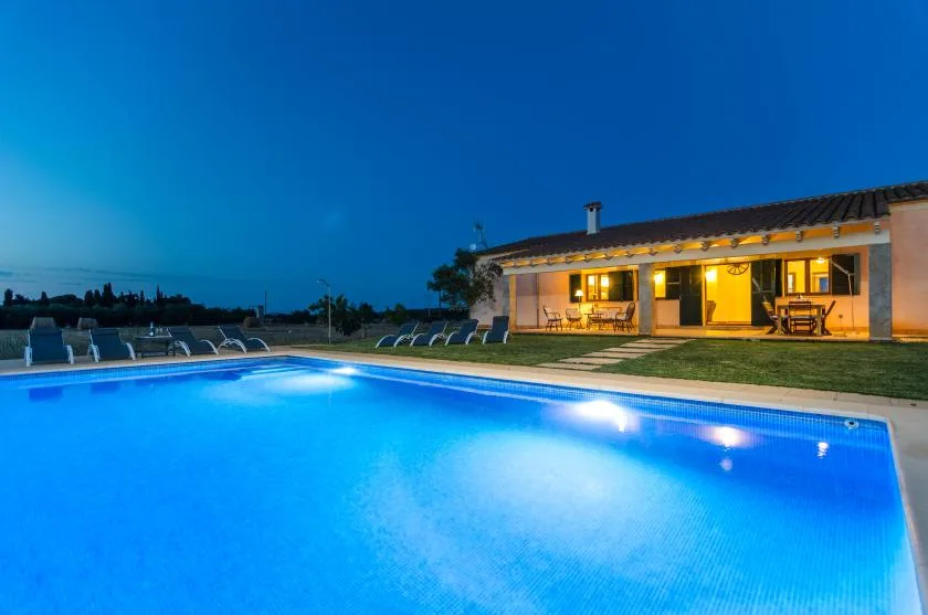 Una piscina iluminada frente a una casa de una planta al anochecer.