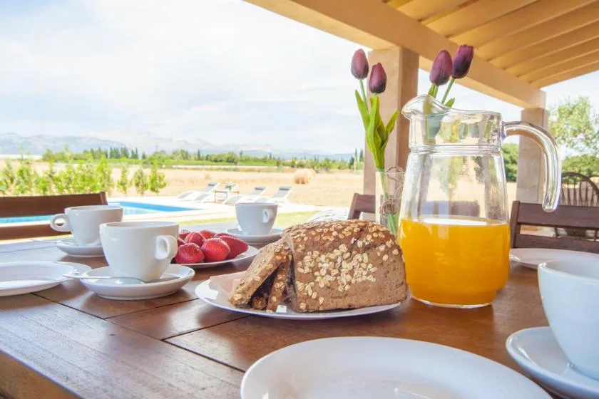 Una mesa de desayuno con pan, tazas, jugo de naranja y flores da a una piscina exterior.