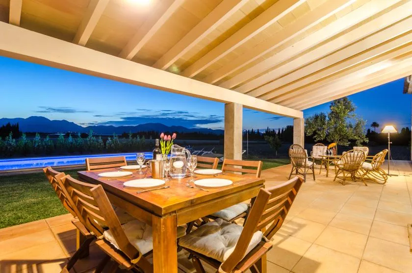 Una zona cubierta de comedor al aire libre, con mesa y sillas de madera, da a una piscina al anochecer.
