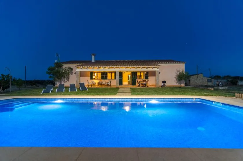 Una casa con un patio iluminado se encuentra detrás de una piscina al anochecer.