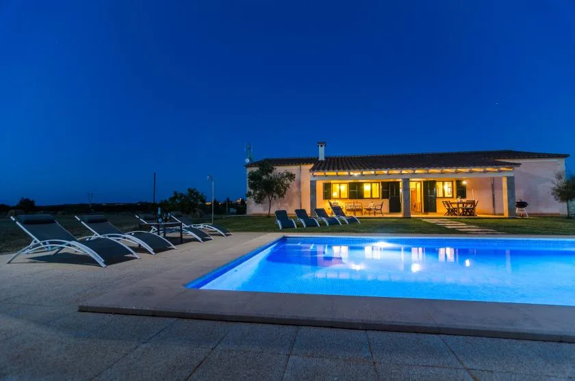 Una casa iluminada está junto a una piscina brillante al anochecer.