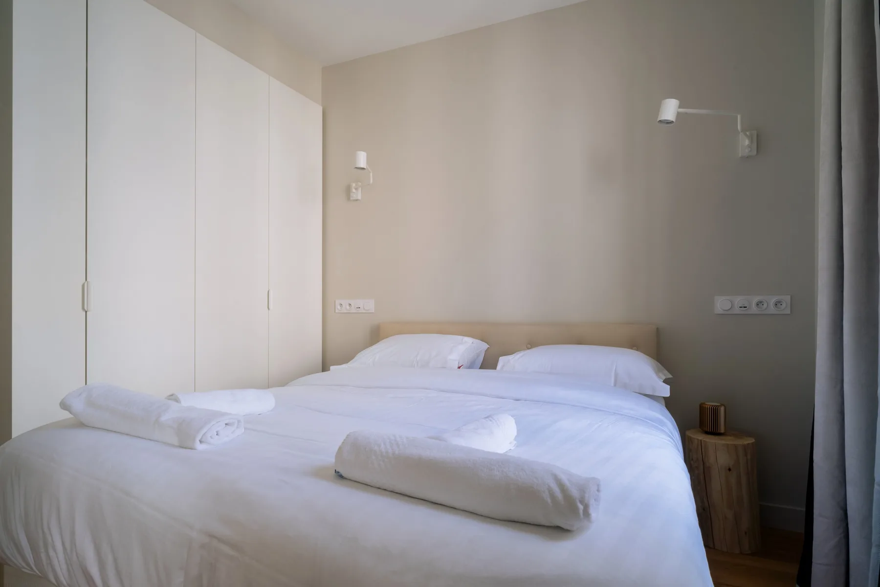 A bedroom with two beds dressed in white linens and wall-mounted lights.