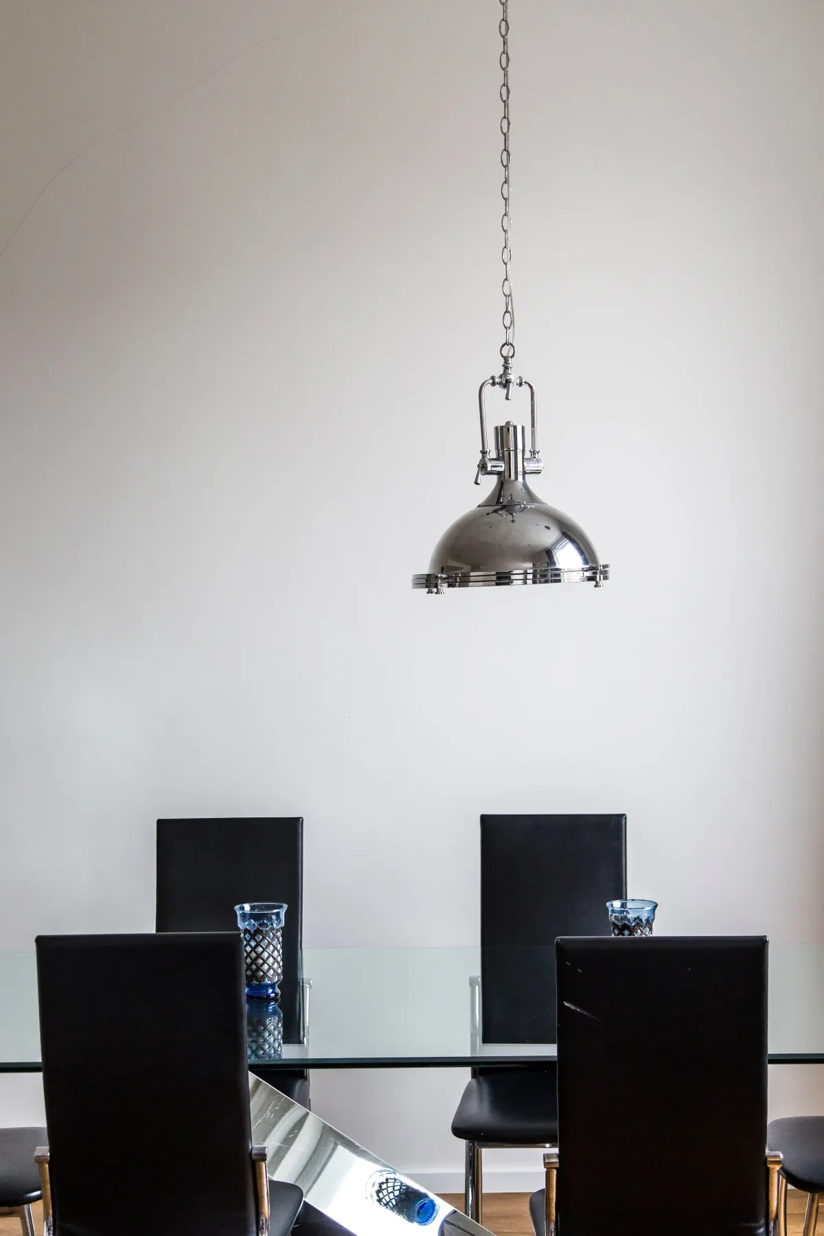 Comedor con mesa de cristal, asientos oscuros y una lámpara metálica colgante.