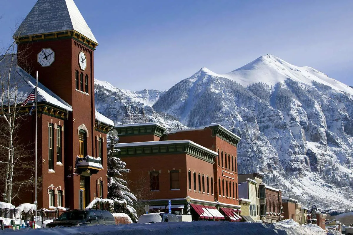 Cumbres nevadas se alzan detrás de una hilera de edificios de ladrillo junto a una calle cubierta de nieve.