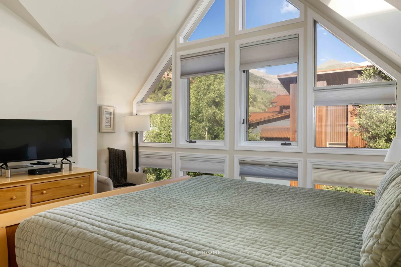 Un dormitorio con una cama grande frente a ventanas altas con vistas a árboles y tejados cercanos.