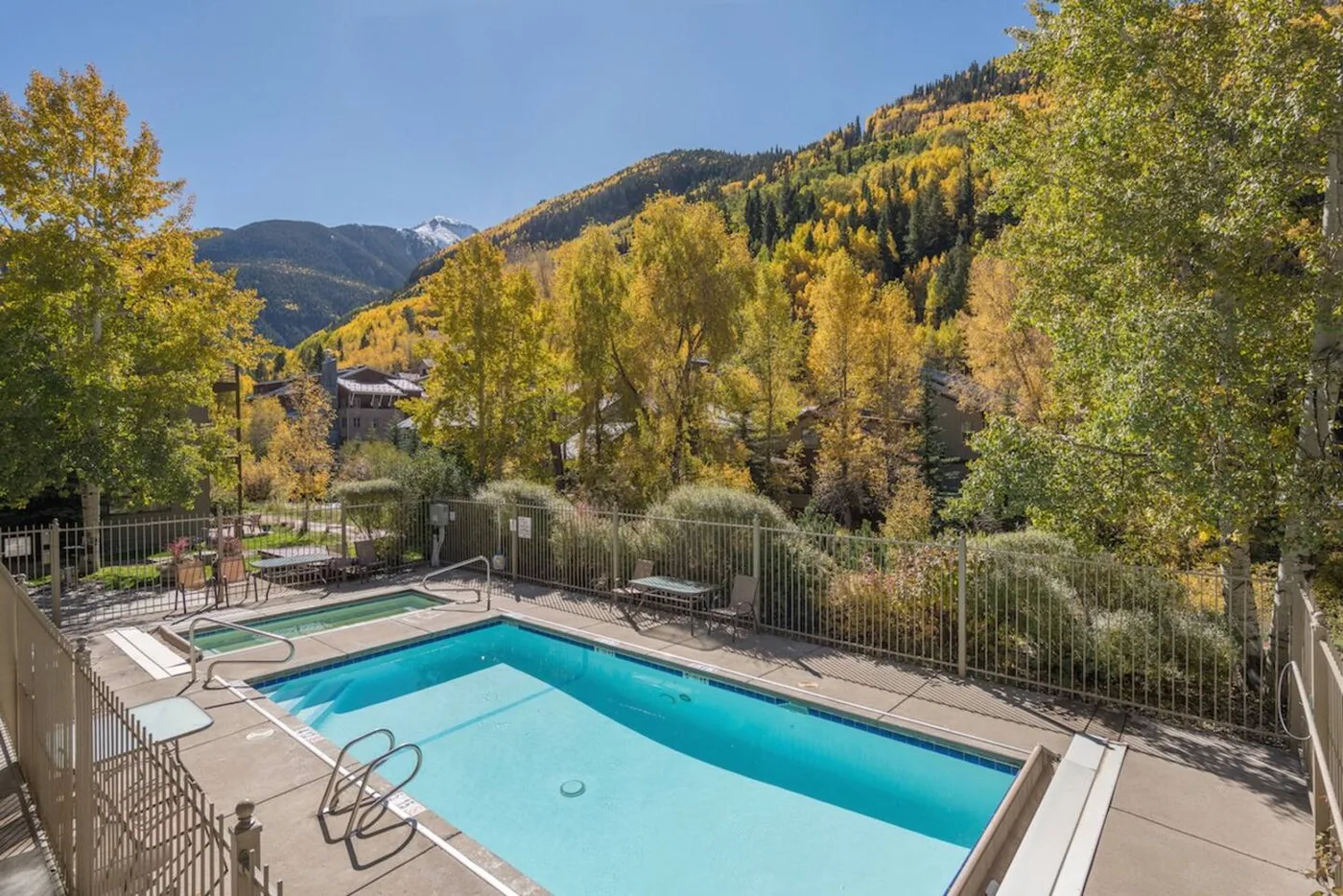 Una piscina al aire libre está junto a una terraza cercada, con árboles otoñales y montañas al fondo.
