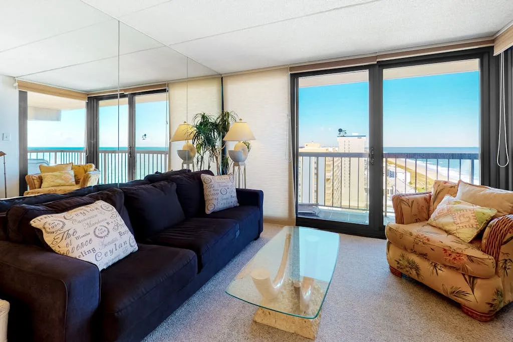 A living room with dark seating, a glass coffee table, and sliding doors overlooking the beach.