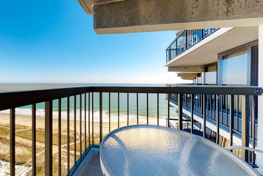 A balcony overlooks the beach and ocean, with an outdoor table in the foreground.