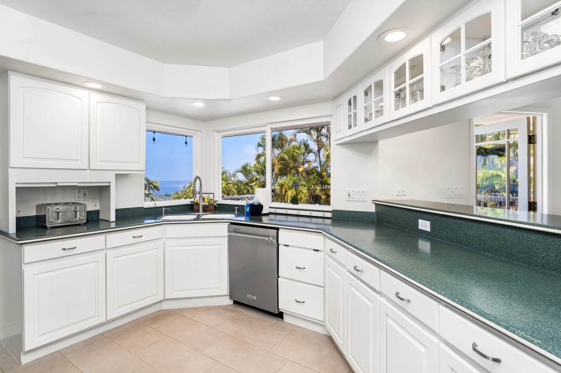 Una cocina luminosa con gabinetes blancos, encimeras verdes y ventanas con vista a palmeras.